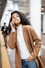 african american woman biting lips while talking on smartphone and looking away in subway on blurred background