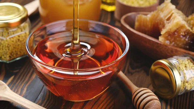 Honey pouring into glass bowl over wooden background with honeycomb and propolis, liquid sugar syrup, flower nectar