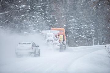 Left-handed driving road maintenance truck clears snow from the road in the forest with a car driving behind it
