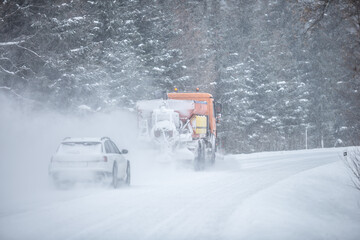 Left-handed driving road maintenance truck clears snow from the road in the forest with a car...