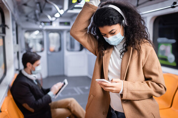 young african american woman in medical mask listening music and using smartphone near man in subway on blurred background