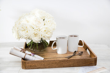A bouquet of white roses in a round glass vase on a table with a cup of tea and a wrapped rolled paper.