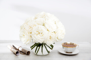 A bouquet of white roses in a round glass vase on a table with a cup of tea and a wrapped rolled paper.