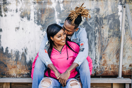 An attractive and fun African-American couple sitting outside on steps in an urban setting with a jacket and denim jeans snuggling and cuddling