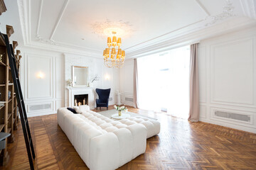 modern interior of a luxurious large bright living room. white expensive sofa and wooden shelving, white walls with moldings and a luxurious chandelier