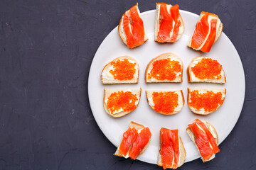 Bruschettes with butter and red caviar and salmon on a white plate on a gray concrete background copy space.