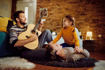 Happy man playing music on acoustic guitar while enjoying with his wife and daughter at home.
