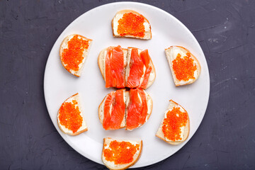 Bruschettes with butter and red caviar and salmon on a white plate on a gray concrete background.