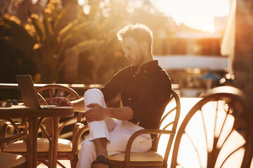 Sits by the table. Young man is outdoors at sunny daytime. Concept of vacation