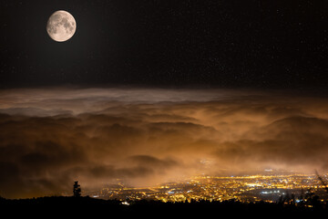Fototapeta na wymiar The full moon shines on the closed cloud cover from above. Under the clouds is the brightly lit town of Puerto de la Cruz on the Spanish island of Tenerife. There are many stars in the sky.