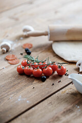 Ripe cherry tomatoes lie on a brown wooden table. Next to the ingredients for making pizza: dough, butter, flour and mushrooms. Natural light. Rustic style