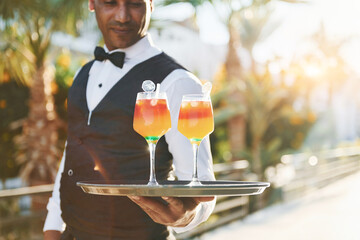 Holds cocktails. Black waiter in formal clothes is at his work outdoors at sunny daytime