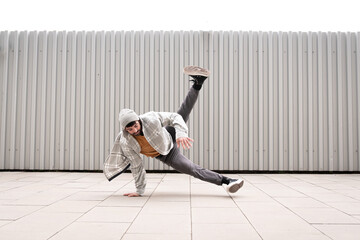 young white caucasian guy with beard gray clothes gray cap and yellow t-shirt dancing break dance doing acrobatics on the floor lifestyle gray metal background fence happy and contented
