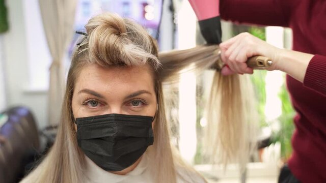 in a beauty salon the hands of a hairdresser dry the blonde hair of a girl in a protective mask with a red brush and a hair dryer. the girl looks at the camera