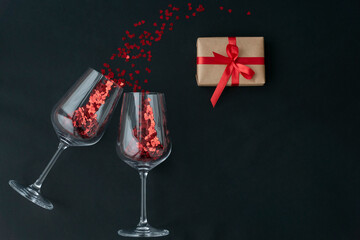 top view of two wine glasses with little red heart sparkling confetti and wrapped giftbox on black background, lots of copy space for text. Love and valentines day concept