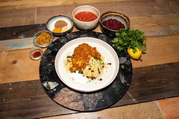 A delicious plate of slow cooked melt in the mouth Chicken Tagine, with couscous surrounded by the ingredients used to make the dish on a rustic wooden kitchen work top.