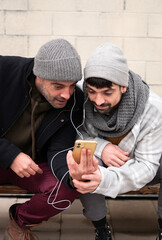 Two adult white caucasian friends wearing warm clothes and scarf listening to music with headphones looking at a yellow smart phone dancing laughing happy lifestyle sitting on a bench
