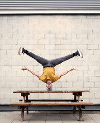 Caucasian boy doing a handstand with his head without hands dancing break dance on top of a bench with jeans and a yellow t-shirt brick background lifestyle