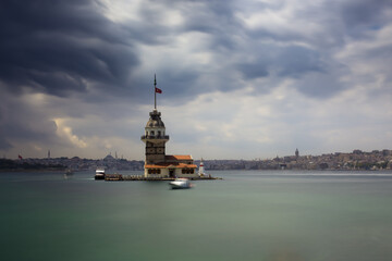 A long exposure photo of a tower looking like a lighthouse on a small island in the sea. Dramatic stormy sky and water are blurred. Oriental city in the background with mosques and minarets.