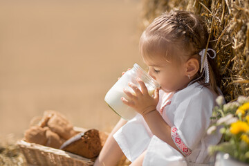 a healthy childhood in harmony with nature. happy little baby in folk clothes sits on a wheat field with a jar of fresh milk.  the sun's rays are reflected in a jar of fresh, tasty milk