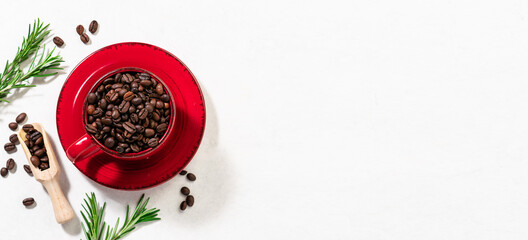 Creative composition with red cup with aromatic coffee beans and bright rosemary twigs on white marble background. Coffee shop banner, cafeteria promo banner. Copy space for your design.