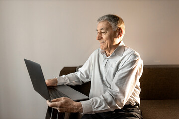 Smiling older man using laptop at home, looking at screen, searching information in internet, happy mature male making video call, sitting on couch.