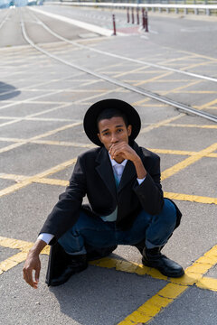 Stylish young man wearing hat crouching on street during sunny day