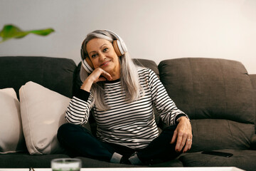 Senior woman wearing headphones smiling while sitting with hand on chin at home