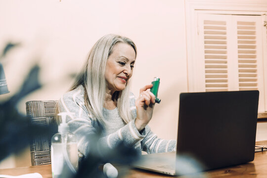 Smiling senior woman showing asthma inhaler during online consultation at home