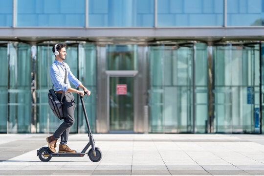 Young businessman wearing headphones riding push scooter on footpath