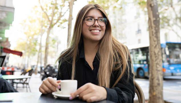 Young woman with coffee cup smiling while sitting at sidewalk cafe in city