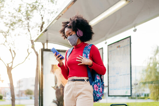 Young woman wearing headphones and protective face mask using smart phone while standing at bus stop