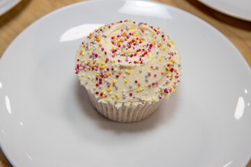 A delicious British cup cake with white icing and multi coloured sprinkles on a white plate