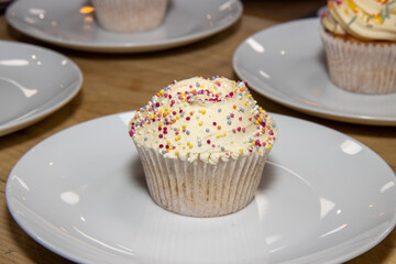 A delicious British cup cake with white icing and multi coloured sprinkles on a white plate
