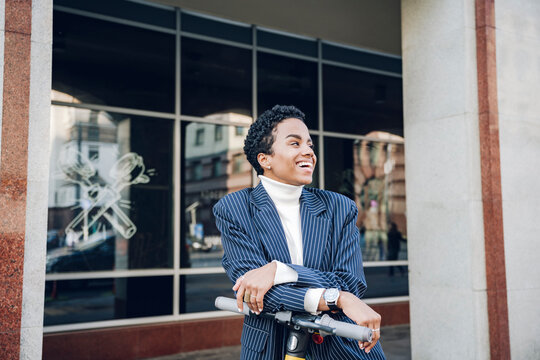 Cheerful businesswoman leaning on handlebar of electric push scooter in city