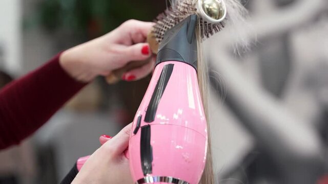 blonde hair is twisted on a round brush and stretched along its entire length, directing air from a pink hairdryer to it
