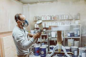 Craftsman wearing protective face mask painting table with spray gun while working at workshop