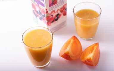 healthy persimon juice in glasses on white with two pieces of fruit at foreground and partially visible cartoon package.