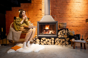 Young woman sitting by a burning fireplace, relaxing with a hot drink in cozy loft style interior...