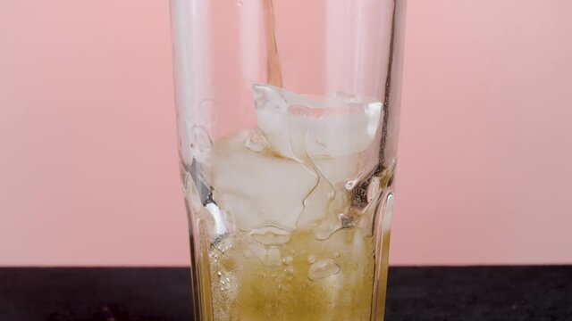 Filling glass with soda water with ice cubes. Action. Close up of pouring soft drink with many bubbles inside the transparent glass isolated on pink background.