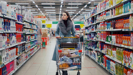 Moscow, Russia - December 29 2020: Young cute girl in a gray coat and a grocery cart with purchases is choosing in the department of household chemicals of a grocery store supermarket