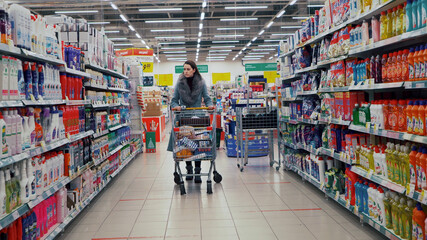Moscow, Russia - December 29 2020: Young cute girl in a gray coat and a grocery cart with purchases is choosing in the department of household chemicals of a grocery store supermarket