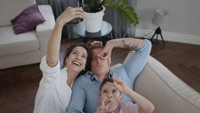 Young Mother Takes Pictures Of Her Family On The Phone. Mom Dad And Son On The Couch. They Show Funny Facial Expressions. Family Having Fun.