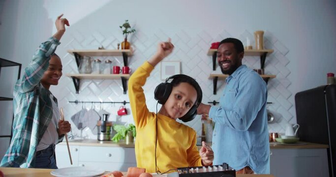 Joyful african family cooking dinner. DJ boy creating funny music on imaginary sound mixer dancing with happy parents in their cozy kitchen. Home party.