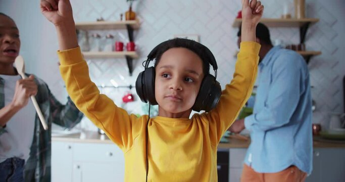 Cute preschool boy wearing headphones acting like DJ having fun with parents dancing into music hanging out at home cooking food.