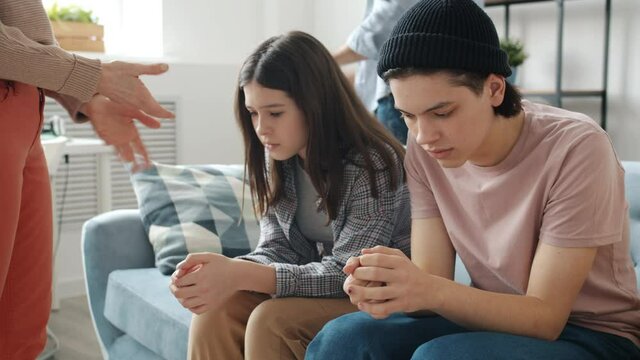 Unhappy teenagers brother and sister are sitting on couch with miserable faces while parents are yelling punishing kids at home. Emotions and childhood concept.