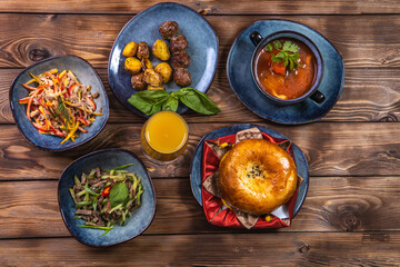 Flat layout of various dishes with sliced vegetables, meat cuts, salads, orange juice and bun on a brown wooden background.