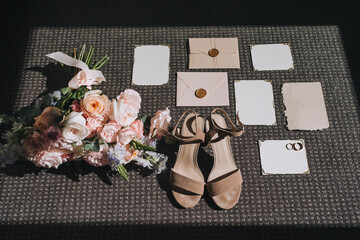 A large and beautiful bouquet of multi-colored roses, beige shoes, bride's sandals, gold rings, paper rugs, invitations lie on the floor with sunlight and shadow. Photography, concept, composition.
