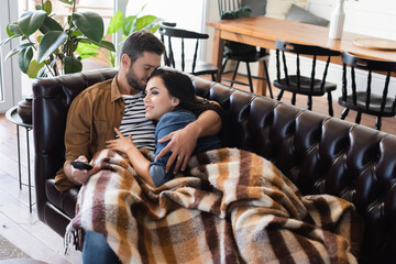young man with closed eyes hugging smiling girlfriend watching tv on leather couch under cozy plaid blanket