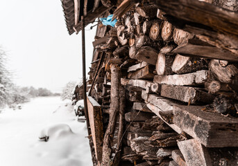 Firewood and boards lie near the wall of the house under a canopy and there is a lot of snow outside